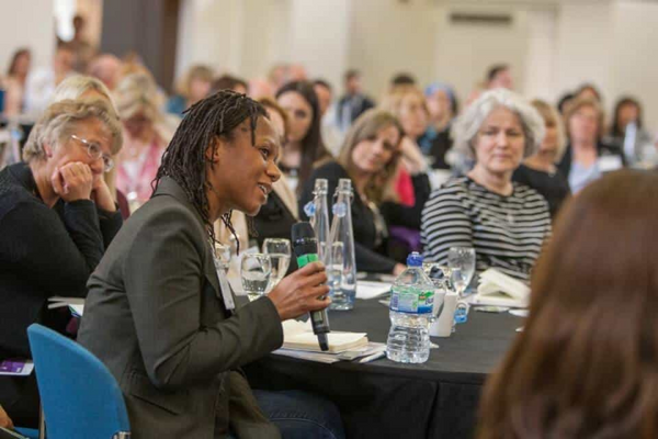 A conference attendee speaks into a microphone as she is sat around a table